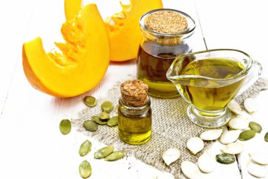 Cam şişede balkabağı yağı, et suyu şişesi ve çuval bezinde bir kavanoz, masada tohumlar, tahta zeminde sebze dilimleri.