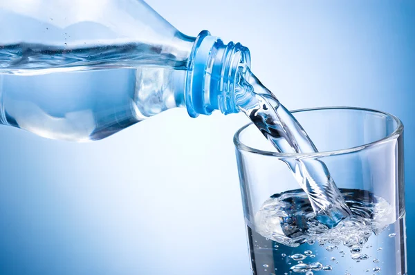 Крупный план заливки воды из бутылки в стекло на синем фоне — стоковое фото