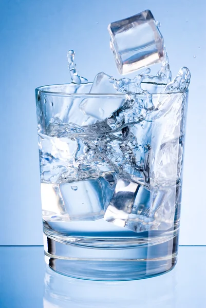 Eiswürfel fallen in ein Glas Wasser auf blauem Hintergrund — Stockfoto