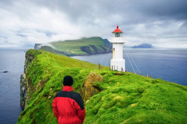 Kırmızı ceketli bir turist Faroe Adaları 'ndaki Holmur Deniz Feneri' nin simgesinin tadını çıkarıyor.
