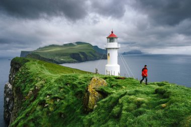 Faroe Adaları 'ndaki Mykines' deki deniz fenerine git. Kırmızı ceketli bir turist yeşil tepeler boyunca yürüyor.