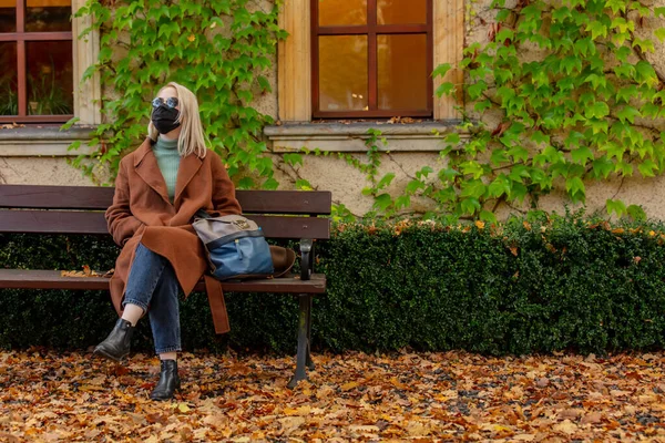 顔のマスクとコートのスタイルブロンドは秋にベンチに座って — ストック写真