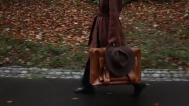 Kahverengi paltolu ve bavullu stil kızı sonbahar parkında yürüyor.