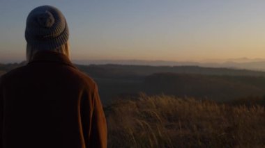 Kız dağlarda bir çayırda yürüyor ve parmaklarıyla çimen dikenlerine dokunuyor.