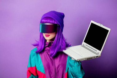 VR gözlüklü ve 80 'lerin eşofmanlı, mor arka planda dizüstü bilgisayarı olan tarz bir kadın.