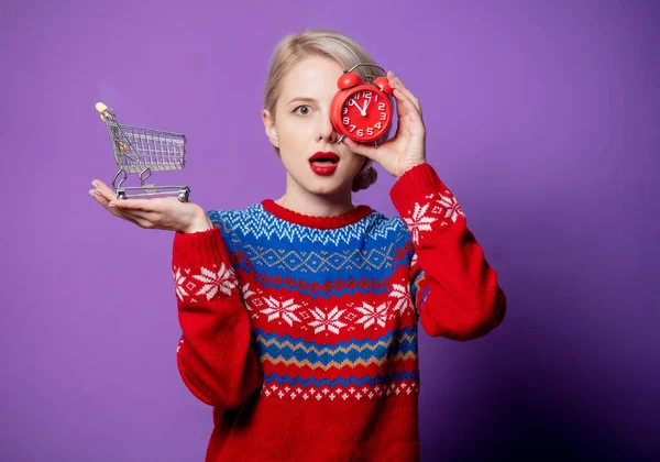 美しい女性でクリスマスセーター目覚まし時計とショッピングカートで紫の背景 — ストック写真