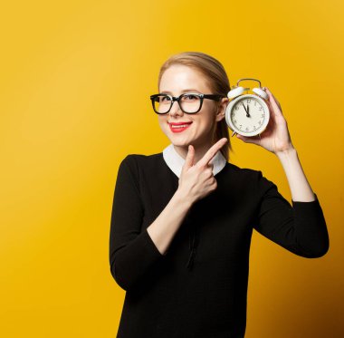 Sarı arka planda çalar saati olan siyah elbiseli, gülümseyen kadın.