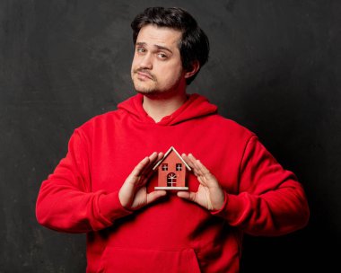 Kırmızı tişörtlü adam karanlık arka planda küçük bir ev tutuyor.