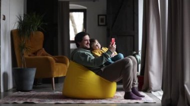 Baba ve oğul evde puf minderde otururken telefon ekranına bakıyorlar.