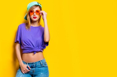 Sarı arka planda turuncu gözlüklü ve şapkalı genç bir kız. 1980 'lerin tarzında kıyafetler.