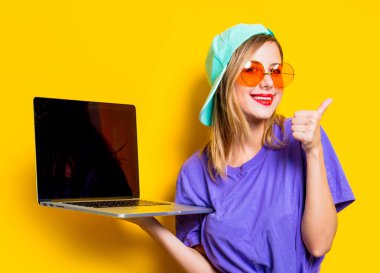 Sarı arka planda turuncu gözlüklü ve dizüstü bilgisayarlı genç bir kız. 1980 'lerin tarzında kıyafetler.