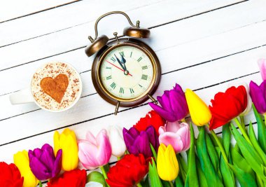 Bir bardak kapuçino, kalp şeklinde bir sembol ve ahşap arka planda çiçeklerin yanında bir saat.