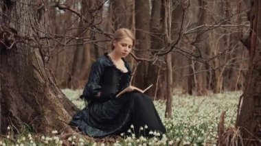 Kitaplı klasik elbiseli kız çayırda oturuyor ormandaki kardelenklerle