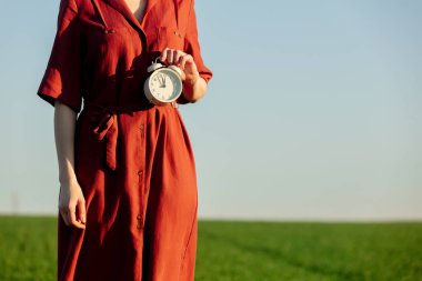 Kırmızı elbiseli kadın, arka planda yeşil buğday tarlasında klasik alarm saati tutuyor.