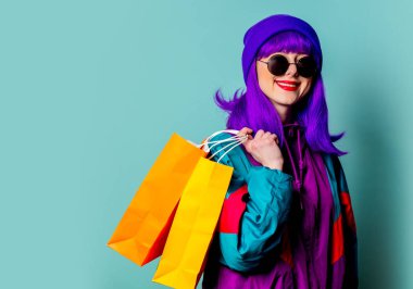 80 'lerin eşofmanlı ve güneş gözlüklü şık alışverişçi kız mavi arka planda alışveriş torbaları tutuyor.