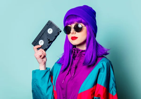 有紫色头发 80年代的运动服和太阳镜的时尚白人女孩拿着蓝色背景的录像带 — 图库照片