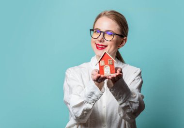 Mavi arka planda küçük bir evi olan beyaz gömlekli güzel sarışının portresi