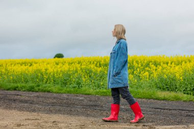 Kot ceketli güzel bir sarışın ve sarı kolza tohumu tarlasında Wellington botu.