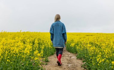 Kot ceketli güzel bir sarışın ve sarı kolza tohumu tarlasında Wellington botu.