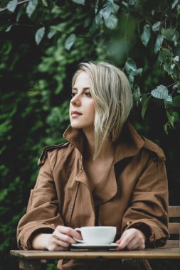 Arka planda ağaçlar olan bir bahçede, ahşap masada bir fincan kahveyle güzel bir kadın.
