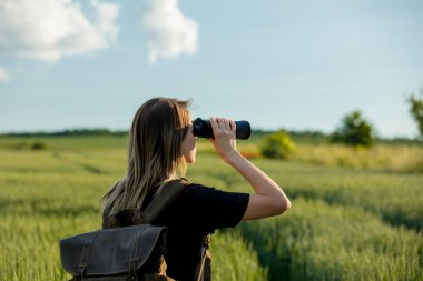 Dürbünlü siyah elbiseli genç kız ve yeşil buğday tarlasında sırt çantası.