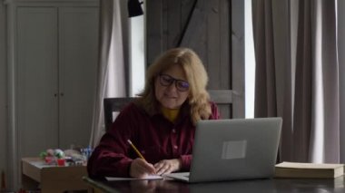 Yaşlı kadın bilgisayarın başına oturur ve kağıda bir şeyler yazar. 