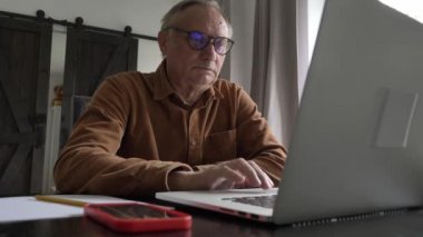 Gözlüklü yaşlı adam evde dizüstü bilgisayar kullanıyor. 4K video