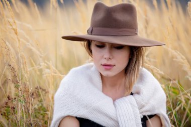 Süveter giyen stil kızı arka planda dağlarla birlikte kırsalda sarı çimenlerde oturuyor.