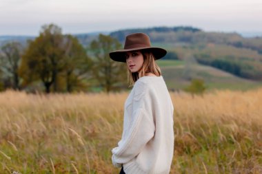 Kırsal kesimde süveter ve şapka giymiş, arkasında dağlar olan bir stil kızı.
