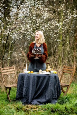 Önlüklü kadın kiraz bahçesinin yanındaki bahçede çay için masayı hazırlıyor.