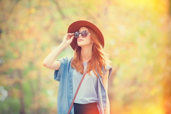 Roodharige meisje in zonnebril en hoed in de herfst park. — Stockfoto