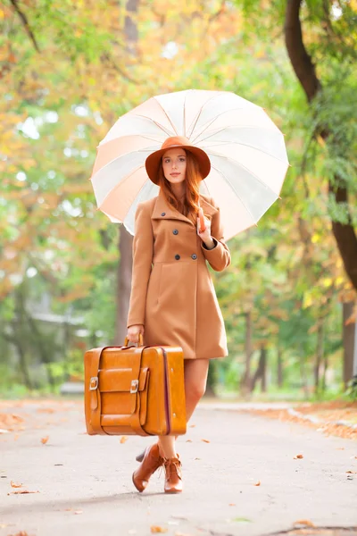 Ragazza rossa con ombrello e valigia nel parco autunnale . — Foto Stock