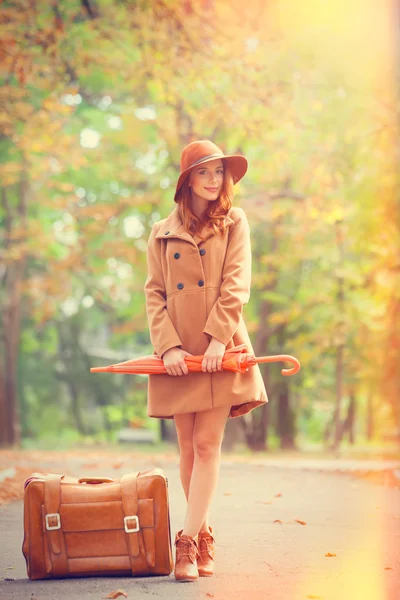 Roodharige meisje met paraplu en koffer in de herfst park. — Stockfoto