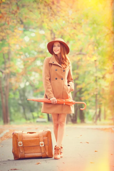 Rothaariges Mädchen mit Regenschirm und Koffer im Herbstpark. — Stockfoto