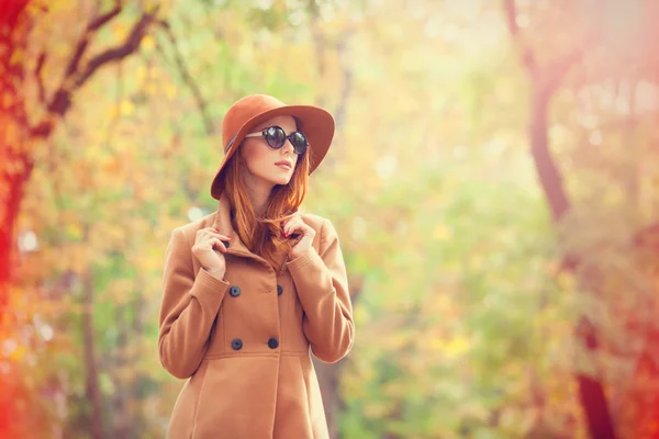 Rödhårig tjej i solglasögon och hatt i höst park. — Stockfoto