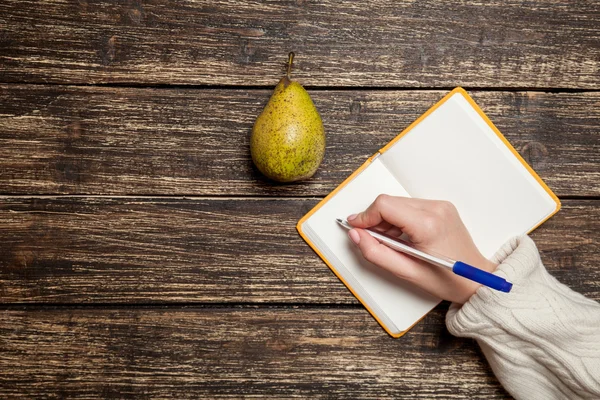 Γυναικείο χέρι που γράφει κάτι στο σημειωματάριο δίπλα στο αχλάδι — Φωτογραφία Αρχείου