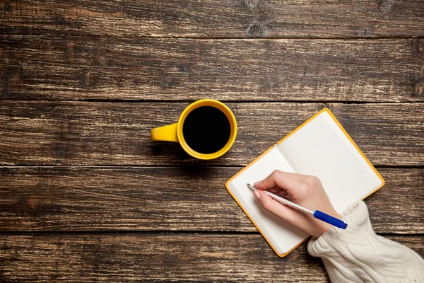 Mujer escribiendo algo a mano en un cuaderno cerca de una taza de café . — Foto de Stock