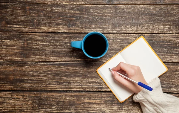 Mujer escribiendo algo a mano en un cuaderno cerca de una taza de café . — Foto de Stock