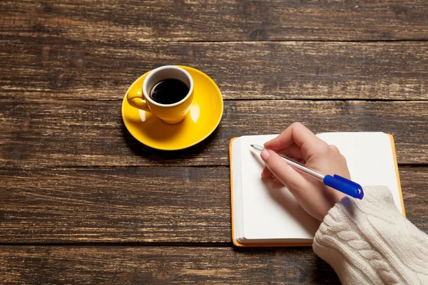 Mujer escribiendo algo a mano en un cuaderno cerca de una taza de café . — Foto de Stock