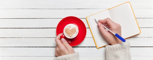 Mão feminina escrevendo algo no caderno perto de xícara de café . — Fotografia de Stock