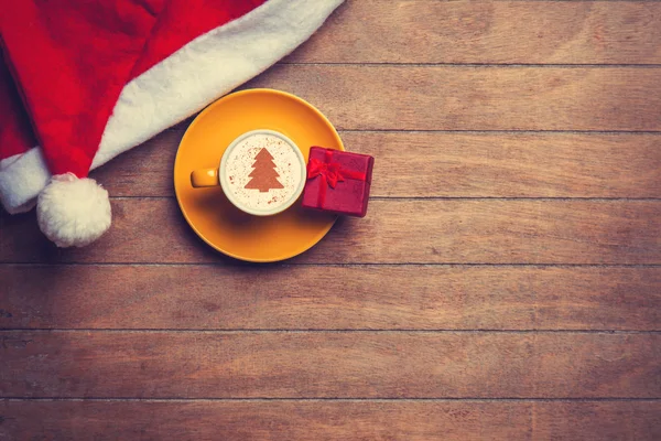 Cappuccino with christmas hat and gift on wooden table. — Stock Photo, Image