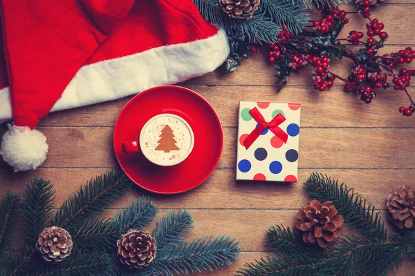 Cup of coffee and gift box with pine and hat on wooden table. — Stock Photo, Image