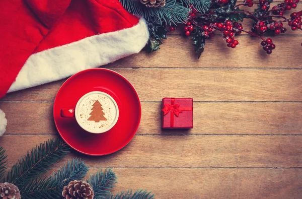 Cup of coffee and gift box with pine and hat on wooden table. — Stock Photo, Image