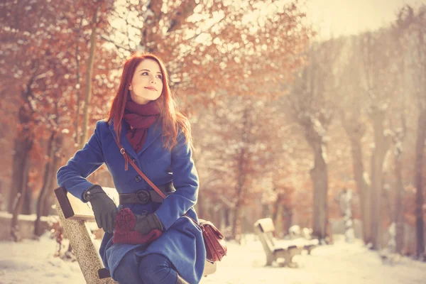 在冬季公园的长椅上坐着的红发女孩. — 图库照片