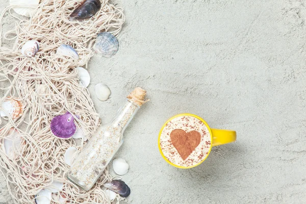 Cup, bottle and net with shells on sand — Stock Photo, Image