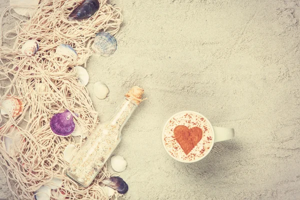 Cup, bottle and net with shells on sand — Stock Photo, Image