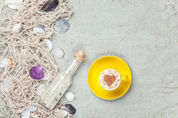 Cup, bottle and net with shells on sand — Stock Photo, Image