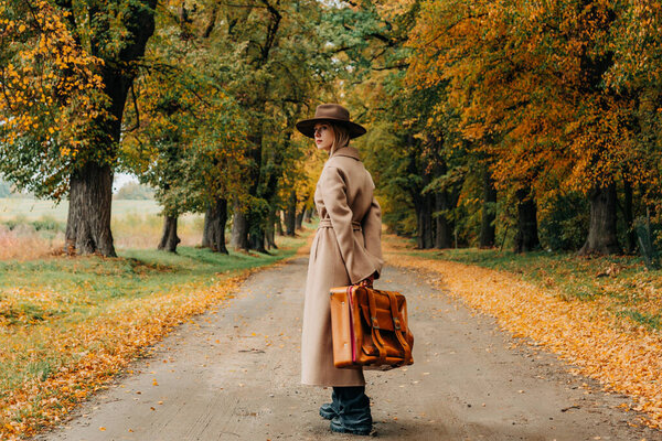 Европейская женщина в коричневом пальто и шляпе 1940-х годов ходит по тропинке через деревья с ярко-желтой осенней листвой
