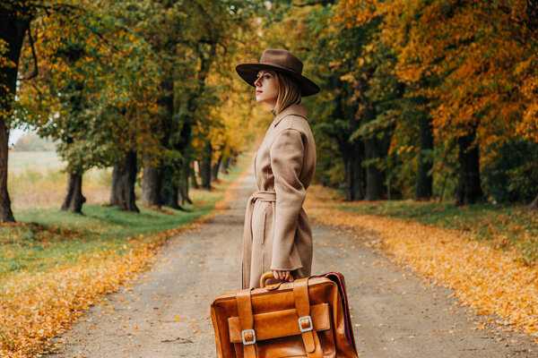 Европейская женщина в коричневом пальто и шляпе 1940-х годов ходит по тропинке через деревья с ярко-желтой осенней листвой