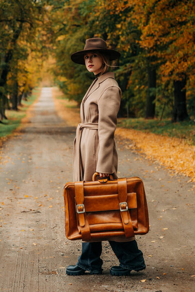 Европейская женщина в коричневом пальто и шляпе 1940-х годов ходит по тропинке через деревья с ярко-желтой осенней листвой